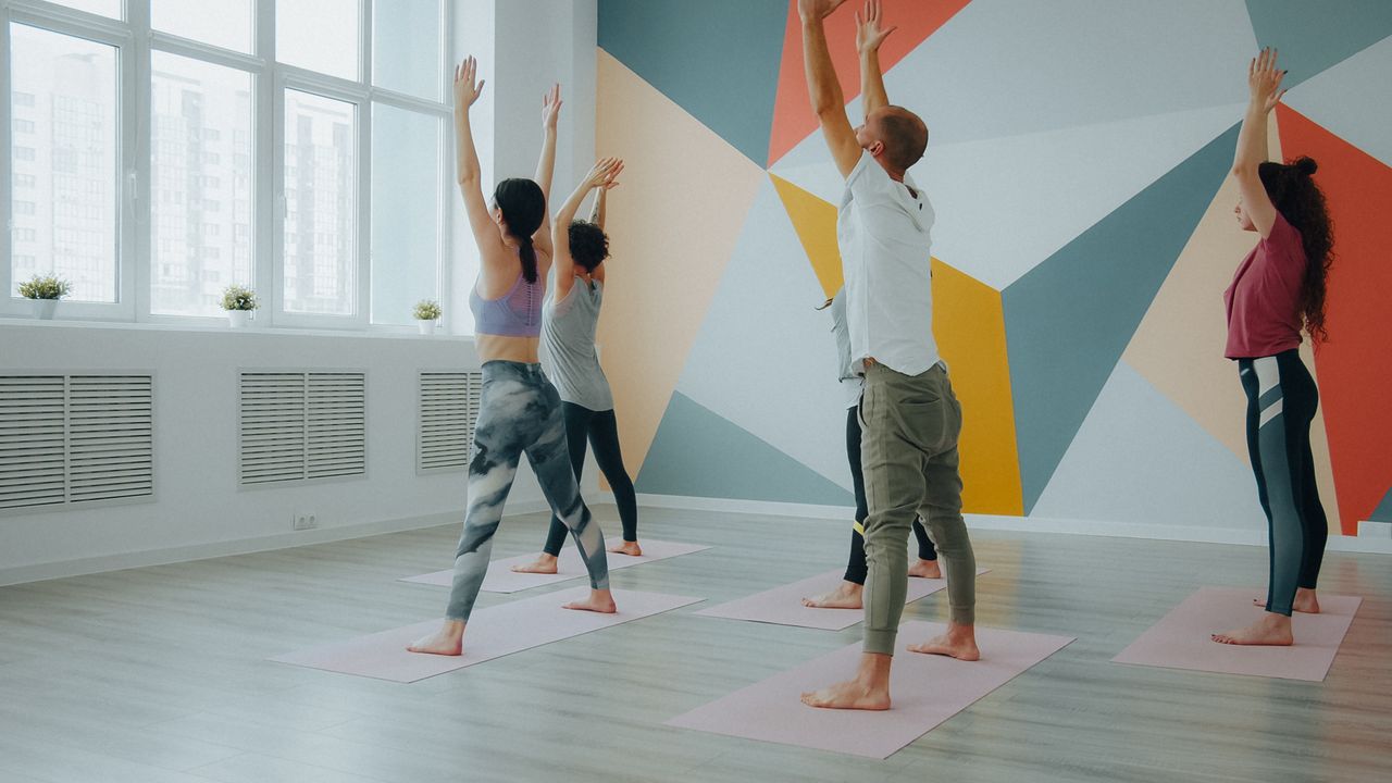 our-approach A diverse group of adults practice yoga in a brightly lit studio with geometric wall design.